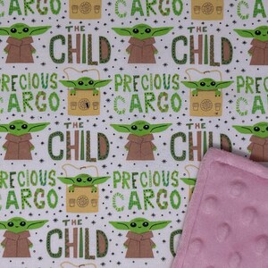 Peut inclure: Tissu blanc avec un motif r&eacute;p&eacute;t&eacute; d'un personnage extraterrestre vert, de robes marron et des mots "THE CHILD" et "PRECIOUS CARGO" en vert. Une couverture rose textur&eacute;e est visible.