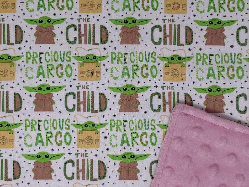 Peut inclure: Tissu blanc avec un motif r&eacute;p&eacute;t&eacute; d'un personnage extraterrestre vert, de robes marron et des mots "THE CHILD" et "PRECIOUS CARGO" en vert. Une couverture rose textur&eacute;e est visible.