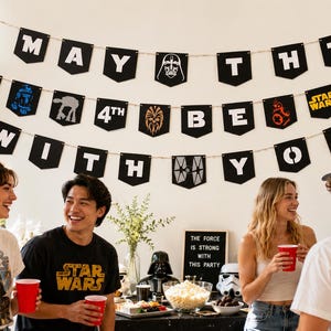 May include: A Star Wars themed party banner featuring black pennants with white letters spelling out May The 4th Be With You, interspersed with character icons including Darth Vader, Chewbacca, and various droids, hung against a white wall.