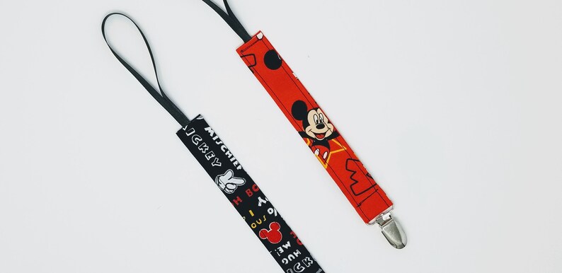 Puede incluir: Dos clips para chupete de beb&eacute;. Uno es rojo con un dise&ntilde;o de Mickey Mouse y un clip plateado. El otro es negro con una mano de Mickey Mouse blanca y texto. Ambos tienen un lazo negro.