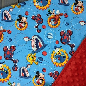 Puede incluir: Una manta para beb&eacute; con una tela azul claro con un patr&oacute;n repetido de Mickey Mouse, Goofy y Donald Duck en un dise&ntilde;o mec&aacute;nico. La manta se combina con una tela de felpa roja texturizada.