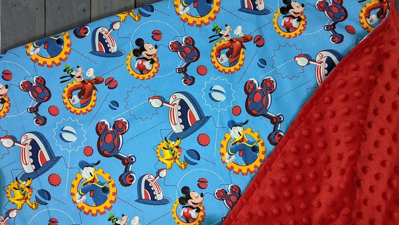 Puede incluir: Una manta para beb&eacute; con una tela azul claro con un patr&oacute;n repetido de Mickey Mouse, Goofy y Donald Duck en un dise&ntilde;o mec&aacute;nico. La manta se combina con una tela de felpa roja texturizada.