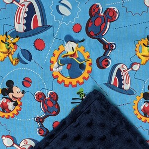 Puede incluir: Una tela azul con un patr&oacute;n repetido de Mickey Mouse, el Pato Donald, Pluto y Goofy en un dise&ntilde;o mec&aacute;nico. Los personajes est&aacute;n sobre un fondo azul claro con acentos rojos, blancos y amarillos. Una manta azul oscuro y texturizada est&aacute; en primer plano.