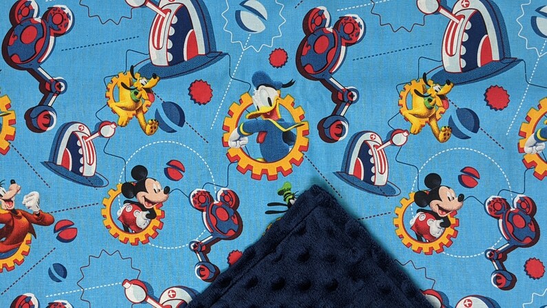 Puede incluir: Una tela azul con un patr&oacute;n repetido de Mickey Mouse, el Pato Donald, Pluto y Goofy en un dise&ntilde;o mec&aacute;nico. Los personajes est&aacute;n sobre un fondo azul claro con acentos rojos, blancos y amarillos. Una manta azul oscuro y texturizada est&aacute; en primer plano.