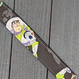 May include: A fabric bookmark featuring Buzz Lightyear from Toy Story. The bookmark has a dark gray background with a repeating pattern and a white ribbon. Buzz Lightyear is depicted in his signature space ranger uniform.