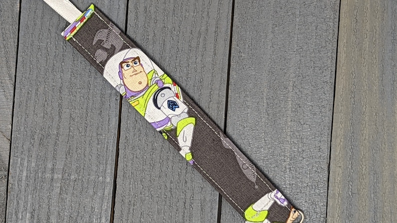 May include: A fabric bookmark featuring Buzz Lightyear from Toy Story. The bookmark has a dark gray background with a repeating pattern and a white ribbon. Buzz Lightyear is depicted in his signature space ranger uniform.