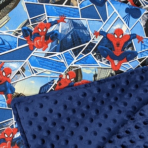 K&ouml;nnte beinhalten: Eine rot-blaue Decke mit Spider-Man-Motiv. Die obere Schicht zeigt ein sich wiederholendes Muster von Spider-Man in verschiedenen Action-Posen vor dem Hintergrund von Stadtlandschaften und blauen geometrischen Formen. Die untere Schicht ist ein marineblauer, strukturierter Stoff.