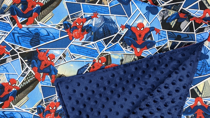 K&ouml;nnte beinhalten: Eine rot-blaue Decke mit Spider-Man-Motiv. Die obere Schicht zeigt ein sich wiederholendes Muster von Spider-Man in verschiedenen Action-Posen vor dem Hintergrund von Stadtlandschaften und blauen geometrischen Formen. Die untere Schicht ist ein marineblauer, strukturierter Stoff.