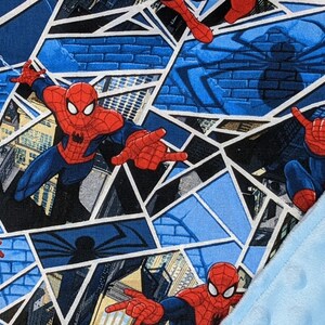 K&ouml;nnte beinhalten: Eine blau-rote Spider-Man-Decke. Das Design zeigt Spider-Man in verschiedenen Action-Posen vor dem Hintergrund von Stadtlandschaften und Spinnennetzmustern. Ein hellblaues, strukturiertes Gewebe ist sichtbar.