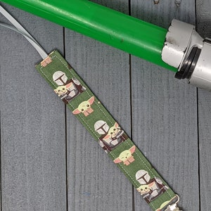 Peut inclure: Marque-page en tissu vert avec un motif r&eacute;p&eacute;t&eacute; de The Mandalorian et Grogu. Un sabre laser vert est en arri&egrave;re-plan. Le marque-page a un ruban blanc attach&eacute;. Le fond est une surface en bois grise.
