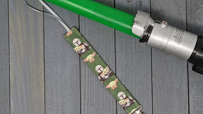 Peut inclure: Marque-page en tissu vert avec un motif r&eacute;p&eacute;t&eacute; de The Mandalorian et Grogu. Un sabre laser vert est en arri&egrave;re-plan. Le marque-page a un ruban blanc attach&eacute;. Le fond est une surface en bois grise.