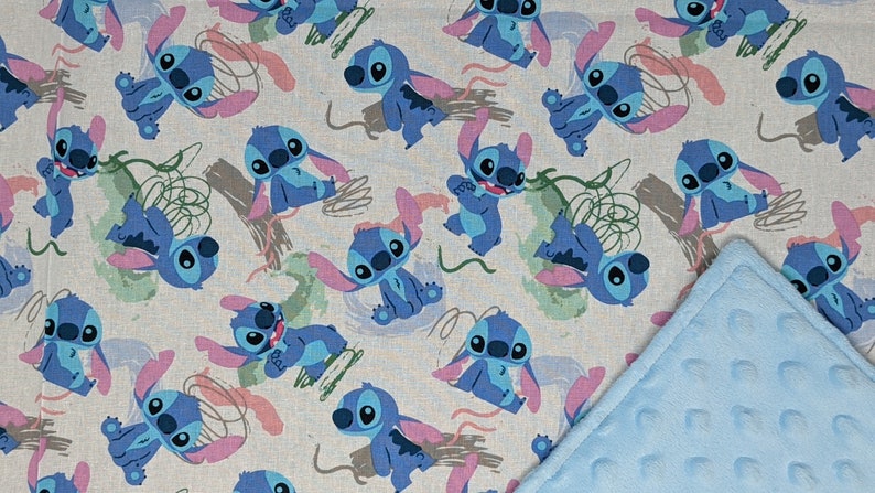 May include: A fabric blanket featuring a repeating pattern of blue Stitch characters on a light gray background, accented with pink ears and green abstract shapes. One corner of the blanket is folded over to reveal a textured, light blue minky fabric.