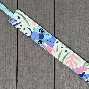 May include: A light blue and green pacifier clip with a floral pattern and a cartoon Stitch character. The clip is attached to a light blue ribbon.