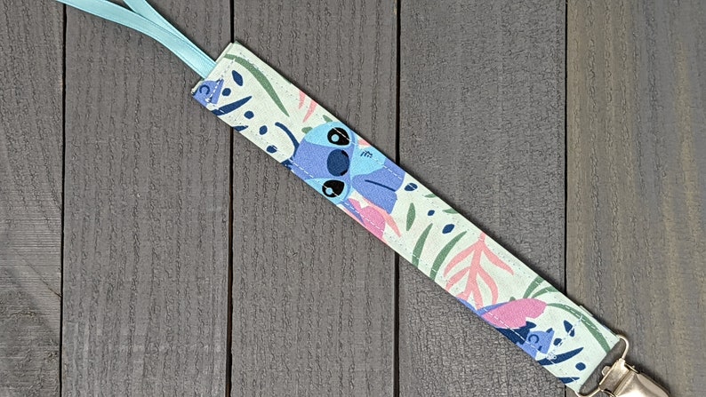 May include: A light blue and green pacifier clip with a floral pattern and a cartoon Stitch character. The clip is attached to a light blue ribbon.