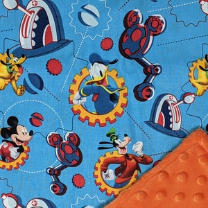 Puede incluir: Tela azul claro con los personajes de dibujos animados Mickey Mouse, Donald Duck, Pluto y Goofy. El dise&ntilde;o incluye engranajes, formas de cabeza de Mickey Mouse y otros elementos mec&aacute;nicos. Se ve una esquina de manta naranja texturizada.