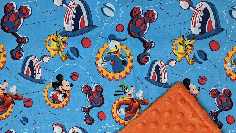 Puede incluir: Tela azul claro con los personajes de dibujos animados Mickey Mouse, Donald Duck, Pluto y Goofy. El dise&ntilde;o incluye engranajes, formas de cabeza de Mickey Mouse y otros elementos mec&aacute;nicos. Se ve una esquina de manta naranja texturizada.