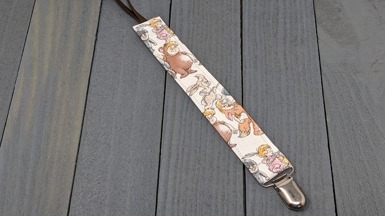 May include: White fabric baby pacifier clip with a brown strap and a silver clip. The fabric features a repeating pattern of cartoon characters from the movie "The Borrowers".