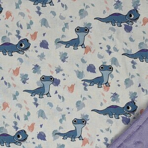 May include: A fabric blanket featuring a repeating pattern of blue cartoon salamanders on a white background with scattered small purple and peach-colored leaves. One corner of the blanket is folded over to reveal a solid light purple textured fabric.