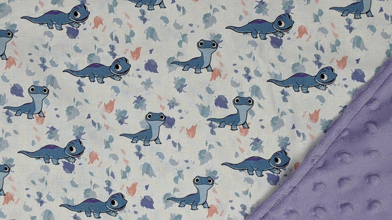 May include: A fabric blanket featuring a repeating pattern of blue cartoon salamanders on a white background with scattered small purple and peach-colored leaves. One corner of the blanket is folded over to reveal a solid light purple textured fabric.