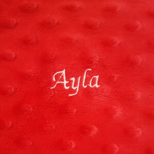 May include: A bright red minky fabric blanket featuring a raised dimpled texture across the surface. The name Ayla is embroidered in white cursive script lettering in the center of the fabric, creating a clear contrast against the red background.