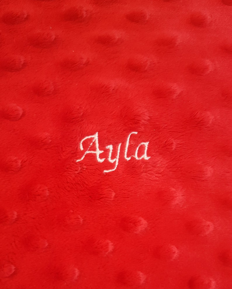 May include: A bright red minky fabric blanket featuring a raised dimpled texture across the surface. The name Ayla is embroidered in white cursive script lettering in the center of the fabric, creating a clear contrast against the red background.