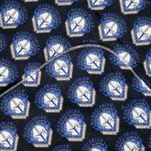 K&ouml;nnte beinhalten: Dunkelblauer Stoff mit einem sich wiederholenden Muster eines Mandalorianer-Helm-Designs in Wei&szlig;, Blau und Grau. Der Text "MANDALORIAN HUNTER BOUNTY" ist ebenfalls sichtbar. Der Stoff ist gefaltet und zeigt eine wei&szlig;e Unterseite.