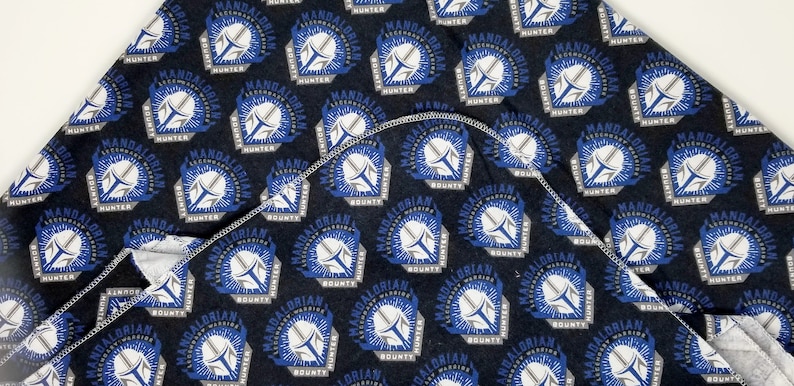 K&ouml;nnte beinhalten: Dunkelblauer Stoff mit einem sich wiederholenden Muster eines Mandalorianer-Helm-Designs in Wei&szlig;, Blau und Grau. Der Text "MANDALORIAN HUNTER BOUNTY" ist ebenfalls sichtbar. Der Stoff ist gefaltet und zeigt eine wei&szlig;e Unterseite.