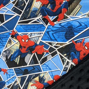 K&ouml;nnte beinhalten: Ein Stoff mit einem sich wiederholenden Muster von Spider-Man in Actionposen. Das Design beinhaltet blaue, rote und wei&szlig;e Farben, mit einem geometrischen Hintergrund und einem schwarzen, strukturierten Stoff auf der rechten Seite.