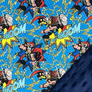 May include: A blue blanket featuring a comic book design with the character Thor, lightning bolts, and the word "THOOM". The blanket has a navy blue, textured backing.