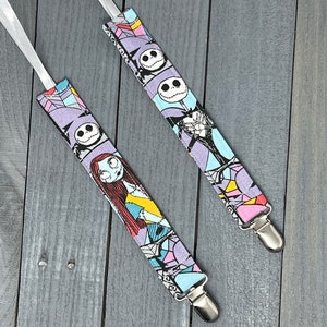 May include: Two pairs of fabric baby suspenders with a Nightmare Before Christmas print. The suspenders are purple, blue, pink, and white with images of Jack Skellington and Sally. The suspenders have silver clips.