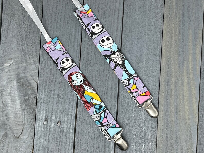 May include: Two pairs of fabric baby suspenders with a Nightmare Before Christmas print. The suspenders are purple, blue, pink, and white with images of Jack Skellington and Sally. The suspenders have silver clips.