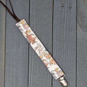 May include: A white fabric pacifier clip with a brown strap and a silver clip. The fabric features a repeating pattern of cartoon characters.