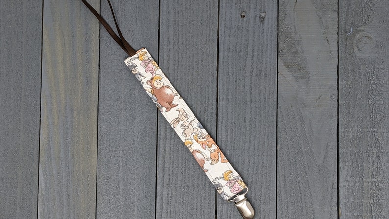 May include: A white fabric pacifier clip with a brown strap and a silver clip. The fabric features a repeating pattern of cartoon characters.