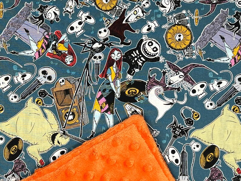 Puede incluir: Una tela azul con un patr&oacute;n repetido de personajes de Pesadilla Antes de Navidad. Los personajes incluyen a Jack Skellington, Sally y Oogie Boogie. La tela est&aacute; cubierta con una manta peluda de color naranja brillante.