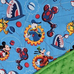 Puede incluir: Una manta para beb&eacute; con un fondo azul con un patr&oacute;n repetido de Mickey Mouse, Donald Duck, Goofy y Pluto. El dise&ntilde;o incluye engranajes y robots con forma de Mickey Mouse. Una tela verde texturizada es visible en la esquina.