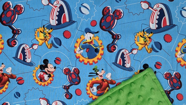 Puede incluir: Una manta para beb&eacute; con un fondo azul con un patr&oacute;n repetido de Mickey Mouse, Donald Duck, Goofy y Pluto. El dise&ntilde;o incluye engranajes y robots con forma de Mickey Mouse. Una tela verde texturizada es visible en la esquina.