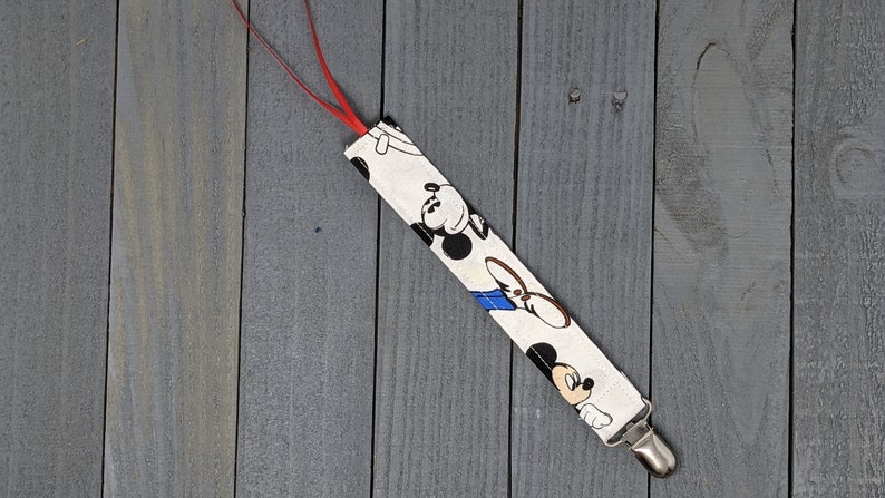 Pu&ograve; includere: Un fermasucchiotto in tessuto bianco con un nastro rosso e una stampa di Topolino. Il fermasucchiotto &egrave; argentato e ha una testa di Topolino all'estremit&agrave;.