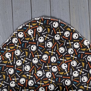 May include: A black fabric with a repeating pattern of white Jack Skellington faces, orange, white, and yellow bones, and gold crossbones. The fabric has a curved edge.