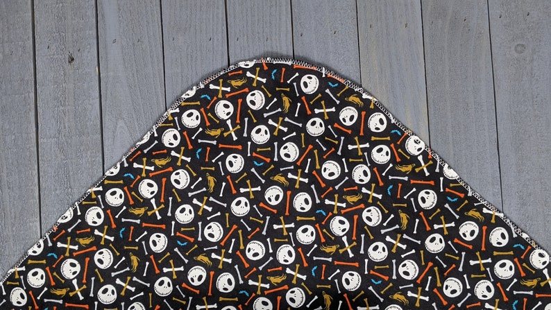 May include: A black fabric with a repeating pattern of white Jack Skellington faces, orange, white, and yellow bones, and gold crossbones. The fabric has a curved edge.