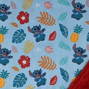 May include: A blue and red fabric baby blanket with a repeating pattern of Stitch from Lilo & Stitch. Stitch is wearing a lei and hula dancing. The pattern also includes pineapples, hibiscus flowers, and tropical leaves. The blanket is reversible with a red minky fabric on the back.