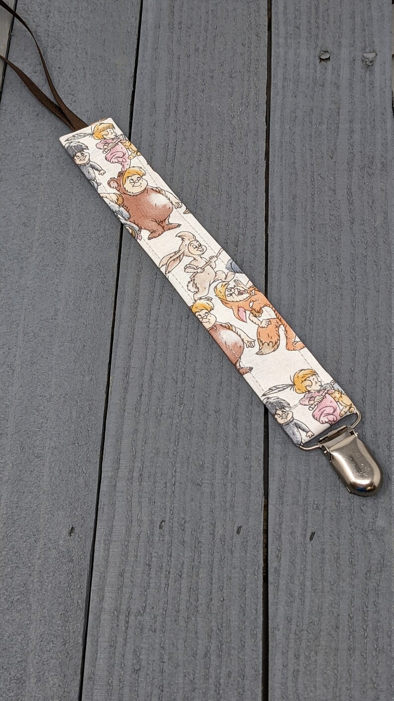 May include: A white fabric baby clip with a brown ribbon and a silver clip. The fabric features a repeating pattern of cartoon characters from the movie "Where the Wild Things Are".