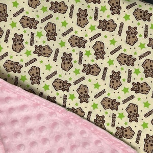 May include: A soft, pink, minky baby blanket with a cream-colored fabric on the other side. The cream fabric has a repeating pattern of a cartoon character with the word "Groot" printed on it.