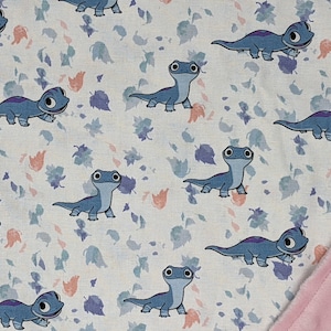 May include: A fabric blanket featuring a repeating pattern of blue cartoon lizards with large eyes and purple back markings, interspersed with small scattered leaves in shades of blue and peach, finished with a textured light pink minky border.
