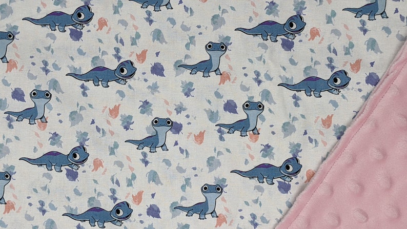 May include: A fabric blanket featuring a repeating pattern of blue cartoon lizards with large eyes and purple back markings, interspersed with small scattered leaves in shades of blue and peach, finished with a textured light pink minky border.