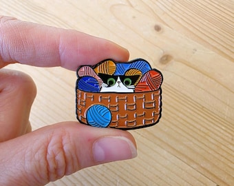 Op de afbeelding: Een kleurrijke emaille pin met een kat met een zonnebril, die uit een bruine mand vol met bollen wol kijkt. De pin heeft een gedetailleerd ontwerp met levendige kleuren, waaronder blauwe, oranje en rode wol.