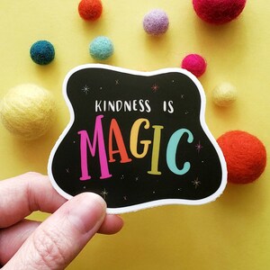 May include: A black sticker with a white border and the text "Kindness is Magic" in rainbow colors. The sticker is on a yellow background with colorful felt balls.