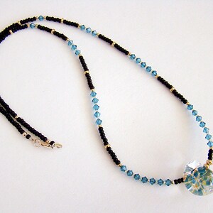 Op de afbeelding: Een zwarte en blauwe kralenketting met een heldere glazen hanger met een bloemenmotief.
