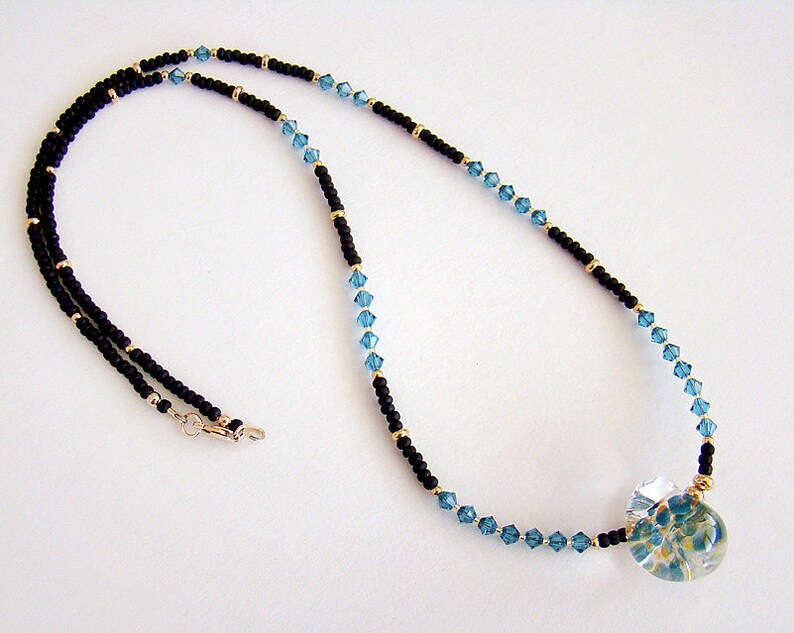Op de afbeelding: Een zwarte en blauwe kralenketting met een heldere glazen hanger met een bloemenmotief.