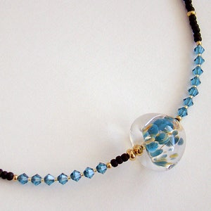 Op de afbeelding: Een delicate ketting met een heldere glazen hanger gevuld met blauwe en gouden vlokken. De ketting is gemaakt met zwarte en blauwe kralen.