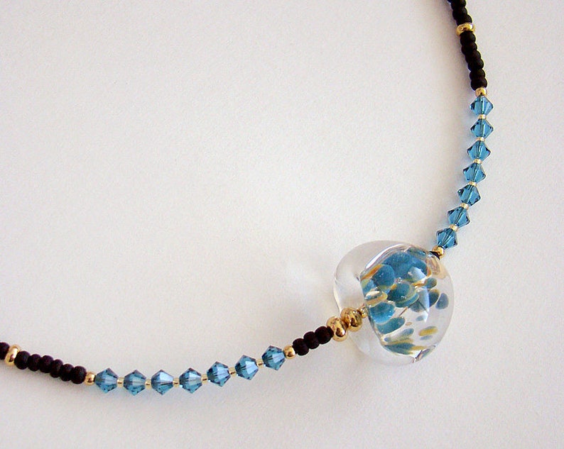 Op de afbeelding: Een delicate ketting met een heldere glazen hanger gevuld met blauwe en gouden vlokken. De ketting is gemaakt met zwarte en blauwe kralen.